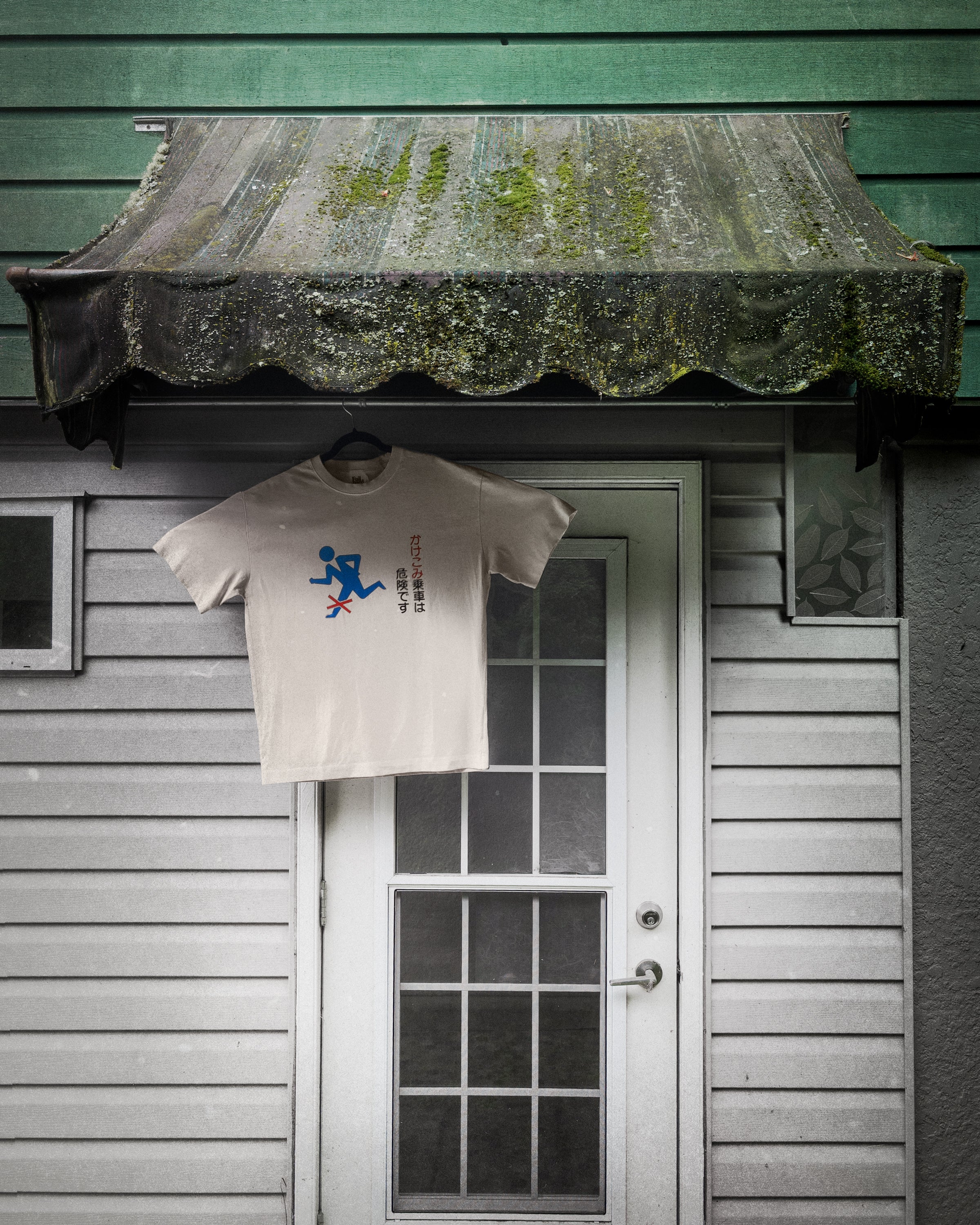 dark background - A white graphic t-shirt hangs from a black hanger on the doorknob of a white wooden door, set against a rustic green and gray house facade with a mossy awning.